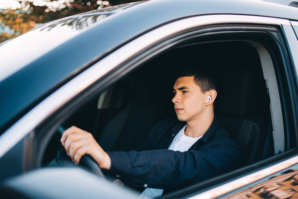 A young guy behind the wheel of a car will use wireless headphones. - Zdjęcie, obraz