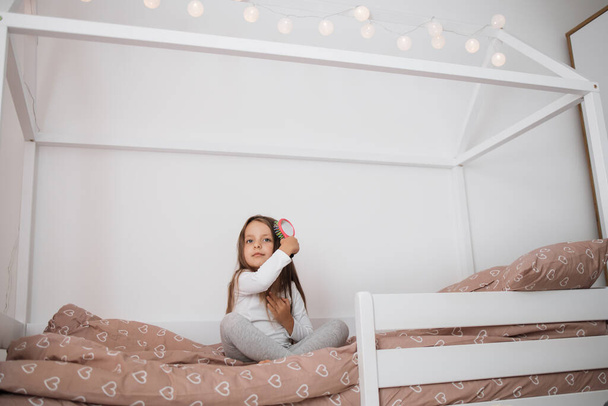 5 years old girl brushing her long hair in her bedroom in the morning. Funny little female child combing her hair. Happy baby sitting on the bed. - Foto, Imagem