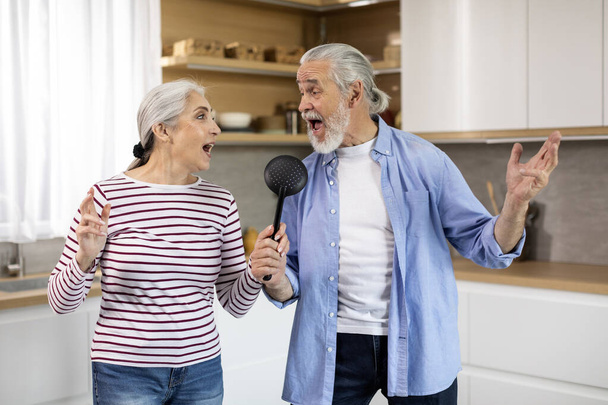 Счастливой пенсии. Радостные пожилые супруги веселятся вместе дома, веселый старший муж и жена дурачатся в интерьере кухни, поют и смеются, используя лопатку как микрофон, копируют пространство - Фото, изображение