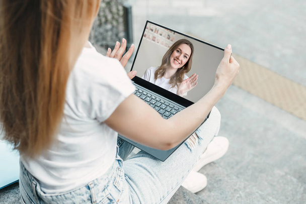 A student girl sitting with laptop on the stairs and making a video call to her friend. Caucasian girl waving from a laptop screen. Video calls, video communication concept - Foto, Imagem