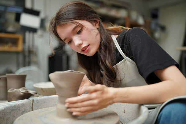 Primo piano di concentrato bella artigiana in grembiule seduto alla ruota della ceramica e utilizzando strumento artigianale mentre modellare vaso di argilla bagnata - Foto, immagini