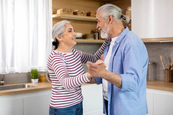 Happy Spouses. Loving Senior Husband And Wife Dancing In Kitchen Interior, Cheerful Elderly Man And Woman Having Fun Together At Home, Holding Hands And Smiling To Each Other, Free Space - Zdjęcie, obraz