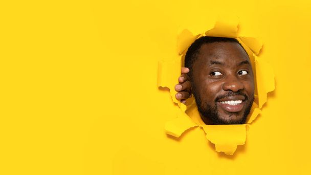 Joyful black man looking aside at free copy space, standing in torn yellow paper hole, noticing funny pleasant unexpected offer, space for text or design - Photo, Image