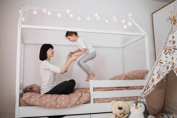 Cheerful mum babysitter play with cute active small kid girl jump on bed, happy carefree mother and little child daughter holding hands laughing having fun feel joy at home in modern bedroom interior - Photo, image