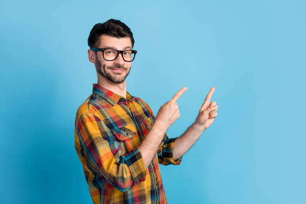 Portrait of young cheerful optimistic handsome man in eyewear dressed plaid shirt directing empty space isolated on blue color background. - Fotografie, Obrázek