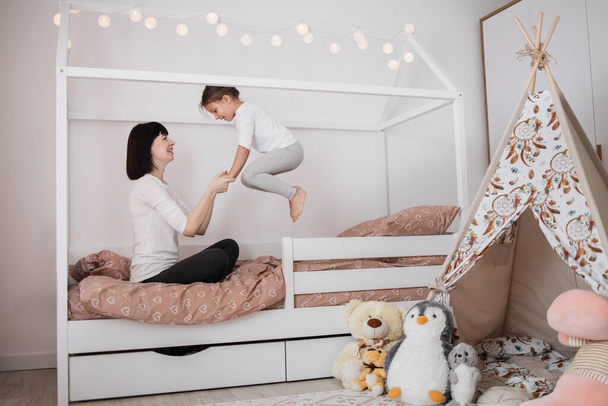 Cheerful mum babysitter play with cute active small kid girl jump on bed, happy carefree mother and little child daughter holding hands laughing having fun feel joy at home in modern bedroom interior - Foto, afbeelding