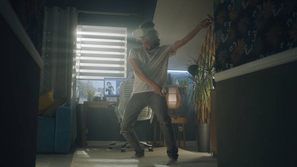 Guy moving while playing video game at home using VR headset and wireless controllers in spare time. Virtual reality device. Cyberspace and metaverse - Fotoğraf, Görsel