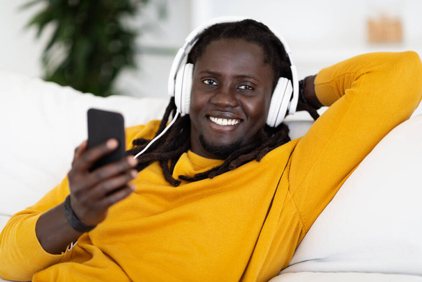 Όμορφος νεαρός μαύρος τύπος με Smartphone και ακουστικά χαλαρώνοντας στον καναπέ στο σπίτι, χαμογελώντας Αφροαμερικανός άνδρας με Dreadlocks ακούγοντας αγαπημένη μουσική σε απευθείας σύνδεση, Πορτρέτο closeup με ελεύθερο χώρο - Φωτογραφία, εικόνα