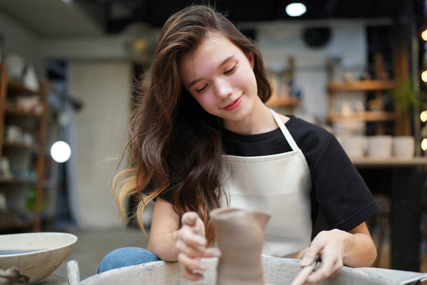 Primo piano di concentrato bella artigiana in grembiule seduto alla ruota della ceramica e utilizzando strumento artigianale mentre modellare vaso di argilla bagnata - Foto, immagini