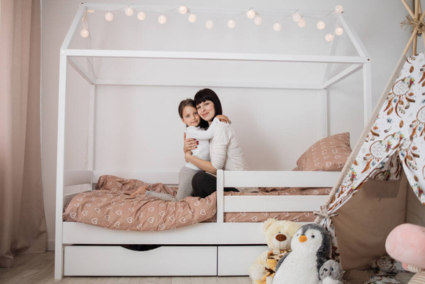 A nice girl and her mother hugging enjoying sunday morning. Good time at home. Child wakes up from sleep. Family playing on the bed in the bedroom. - Фото, изображение