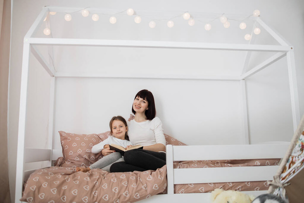 Portrait of cheerful caucasian preschooler girl and her young lady mother reading funny interesting children book together while sitting on bed in bedroom interior. - Foto, Bild