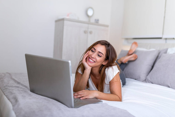 Uśmiechnięta kobieta leżąca na łóżku przed laptopem z podniesionymi nogami. Happy casual piękna kobieta pracuje na laptopie siedzi na łóżku w domu. Wolny strzelec pracujący w domu - Zdjęcie, obraz