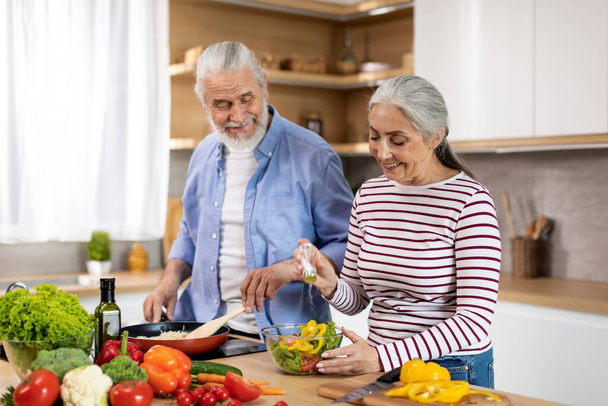 Готовим вместе. Счастливые старшие супруги готовят обед на кухне, веселая пожилая пара готовит еду дома, улыбающийся пожилой джентльмен перемешивает еду со шпателем, в то время как его жена приправляет салат - Фото, изображение