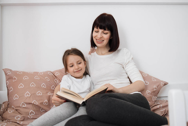 Close up of smiling mom and little daughter relax in cozy bed read funny interesting children book together, happy mother and small preschooler girl child enjoy fairytale rest in comfortable bedroom - Фото, зображення