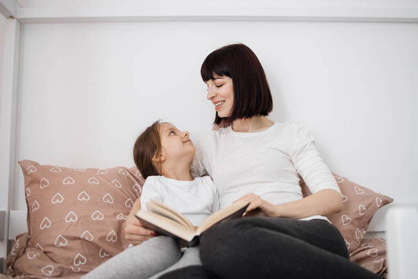 Close up of smiling mom and little daughter relax in cozy bed read funny interesting children book together, happy mother and small preschooler girl child enjoy fairytale rest in comfortable bedroom - Fotografie, Obrázek
