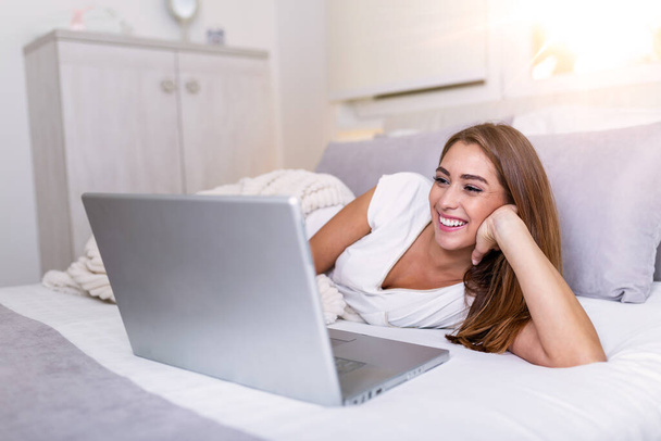 ラップトップコンピュータを使用してベッドでリラックスした美しい女性。笑顔でベッドの上に寝そべっている美しい女性は、午前中にマンションでノートパソコンで働いています。家で働いてるフリーランスの女の子 - 写真・画像