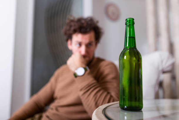 Θλιβερός νεαρός εθισμένος άντρας που αισθάνεται άσχημα πίνοντας μπύρα μόνος στο σπίτι, στρεσαρισμένος μοναχικός αλκοολικός που πάσχει από εθισμό στο αλκοόλ που έχει πρόβλημα, έννοια αλκοολισμού - Φωτογραφία, εικόνα