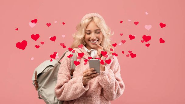 Onnellinen naisopiskelija kaunis nuori blondi nainen käyttää älypuhelinta, kutoma poikaystävänsä verkossa seisoessaan reppu vaaleanpunainen tausta, punainen sydän ympärillä, studio laukaus, kollaasi - Valokuva, kuva