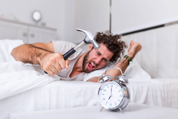 El joven intenta romper el despertador con un martillo, Destruir el reloj. Hombre acostado en la cama apagando un despertador con martillo en la mañana a las 7 am. - Foto, imagen