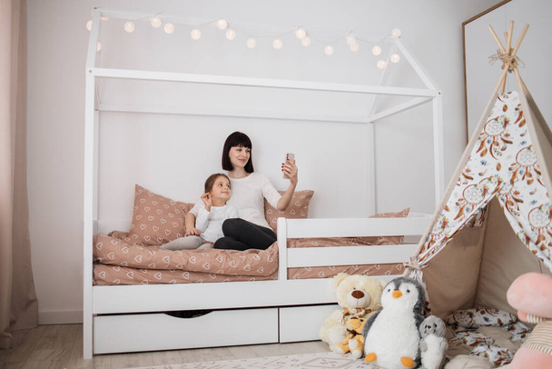 Young mother and her daughter girl are playing in bedroom. Funny mom and lovely child having video call using phone sitting in wooden bed. - Foto, Imagem