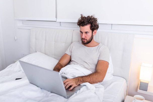 Close-up foto van gefocuste jonge man ondernemer hebben weekend werk gebruik maken van zijn laptop om verslag baas wil liggen op bed in huis binnen. Man die thuis werkt - Foto, afbeelding