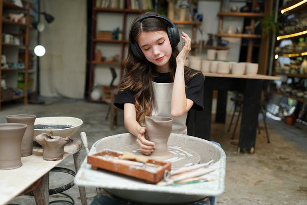 Közelkép a koncentrált gyönyörű kézműves nő kötényben ül a kerámia kerék és a kézműves eszköz, miközben formázása nedves agyag edény - Fotó, kép