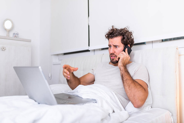 Image of young Caucasian man has morning coffee, sits on bed, working on his laptop and talking on his mobile phone, prepares for examination, uses laptop computer for surfing internet. - Photo, Image