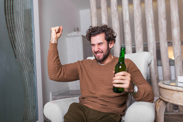 Fanatyczny fan sportu ogląda mecz piłki nożnej w telewizji świętując. Atrakcyjny, szczęśliwy facet oglądający futbol, świętujący zwycięstwo swojej ulubionej drużyny, pijący piwo, siedzący w salonie - Zdjęcie, obraz
