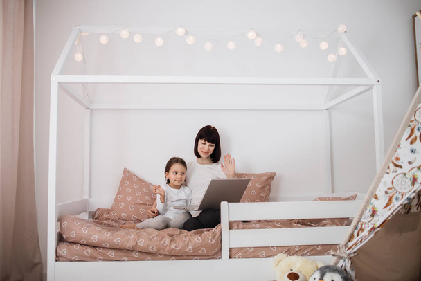 Portrait of caucasian mother with her cute preschool daughter, using laptop having video call with friends sitting on bed in bedroom at home, concept of growth, development, technology. - Фото, изображение