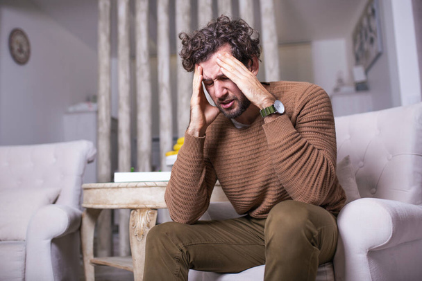 Portret van een knappe man die thuis op een bank zit met hoofdpijn, pijn en een uitdrukking van onwel zijn. - Foto, afbeelding