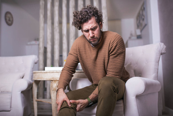 Mladý muž, který trpí bolestí kolena, sedí doma na pohovce. Muž držící se za nohu a trpící bolestí kolena - Fotografie, Obrázek