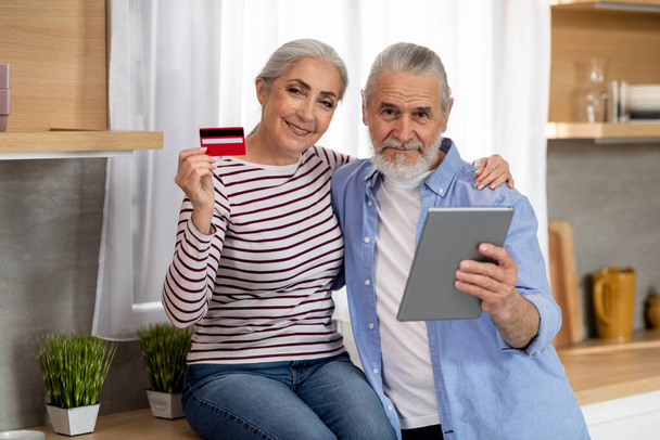 Happy Elderly Couple With Digital Tablet And Credit Card In Hands Posing In Kitchen Interior, Cheerful Senior Husband And Wife Making Online Payments While Relaxing At Home Together, Copy Space - Фото, изображение
