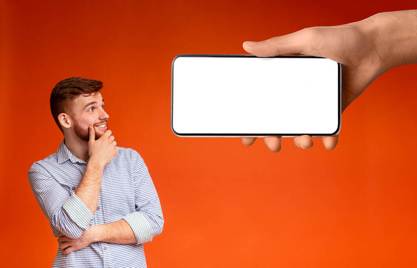 Smiling pensive young caucasian guy with beard looks at huge smartphone with blank screen in hand, isolated on orange background, collage. Choice, great offer, ad, huge sale and new app for work - Фото, изображение