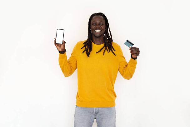 Online-pankkitoiminta. Happy Black Man Holding luottokortti ja tyhjä älypuhelin seisoessaan eristetty yli valkoinen tausta, hymyilevä Afrikkalainen amerikkalainen mies suosittelee App E-Commerce, Mockup - Valokuva, kuva