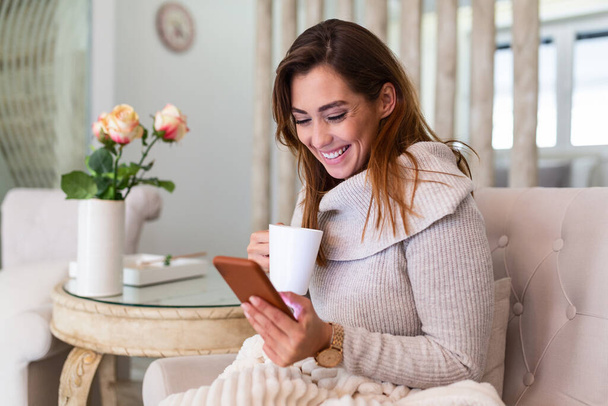 Pretty young woman sitting on sofa and enjoying first morning coffee in cozy apartment. Smiling woman sitting on sofa while drinking hot tea. Happy girl relaxing at home on a bright winter morning. - 写真・画像