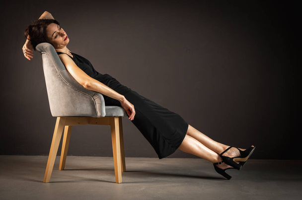 Retrato del estudio de una mujer cansada, torturada, joven y sensual con un vestido negro, sentada en una silla de madera, apoyada en la espalda, sobre un fondo oscuro. Depresión - Foto, Imagen