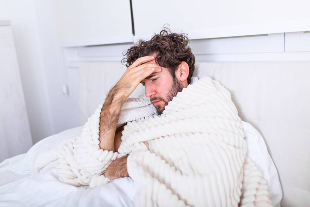 Больной человек с головной болью сидит под одеялом. Больной человек с сезонными инфекциями, гриппом, аллергией лежал в постели. Больной красивый мужчина, покрытый одеялом, лежащий в постели с высокой температурой и гриппом, отдыхающий. - Фото, изображение