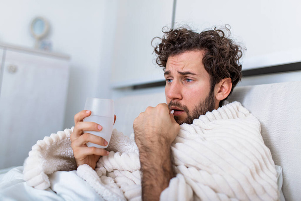 風邪や冬のインフルエンザを患っているパジャマを着てベッドに寝そべっている病気の無駄な男健康管理の概念で薬の錠剤を持っているウイルス水のガラスで薬を飲む - 写真・画像