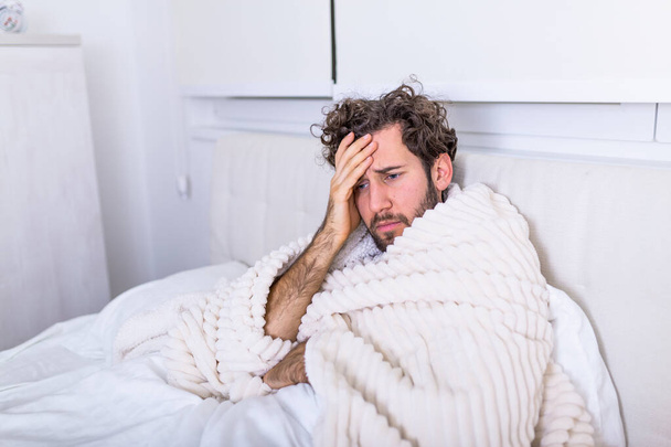 Sick man with headache sitting under the blanket. Sick man with seasonal infections, flu, allergy lying in bed. Sick handsome man covered with a blanket lying in bed with high fever and a flu, resting. - Photo, Image