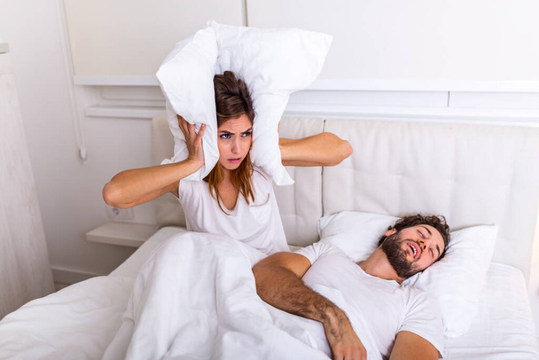 Man snoring while his wife is covering ears with pillow. Woman suffers from her male partner snoring in bed. Noise concept. Real people. Young irritated woman lying in bed with snoring husband - 写真・画像