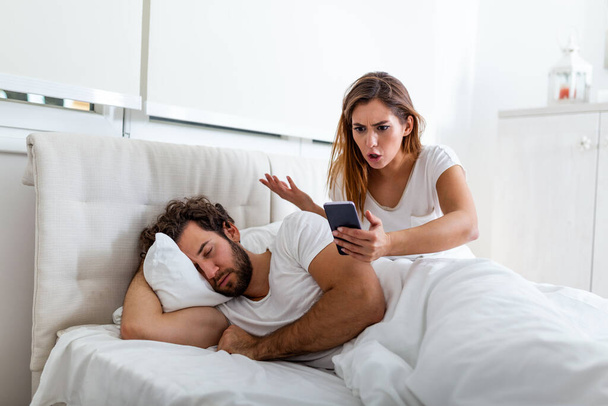 Η ζηλιάρα σύζυγος κατασκοπεύει το τηλέφωνο του συνεργάτη της ενώ αυτός κοιμάται σε ένα κρεβάτι στο σπίτι. Σοκαρισμένη ζηλιάρα γυναίκα κατασκοπεύει το τηλέφωνο του άντρα της ενώ ο άντρας κοιμάται στο κρεβάτι στο σπίτι. - Φωτογραφία, εικόνα