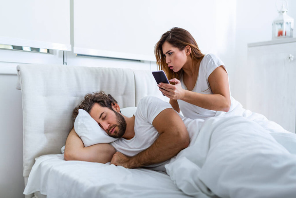 Женщина ревнивая, подозрительная и шпионит в смартфоне своего напарника, пока он спит в спальне. Жена шпионит за телефоном мужа, пока он спит. Концепция недоверия, ревности - Фото, изображение