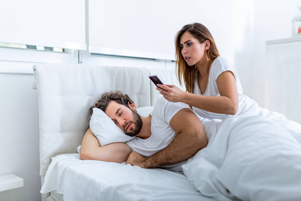 Eifersüchtige Ehefrau spioniert das Telefon ihres Partners aus, während dieser zu Hause in einem Bett schläft. Schockierte eifersüchtige Ehefrau spionierte Handy ihres Mannes aus, während Mann zu Hause im Bett schlief - Foto, Bild