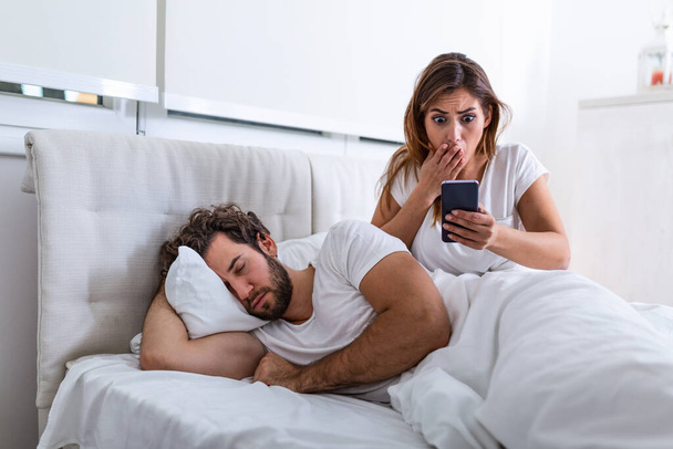 Jealous wife spying the phone of her partner while he is sleeping in a bed at home. Shocked jealous wife spying the phone of her husband while man sleeping in bed at home - Photo, Image