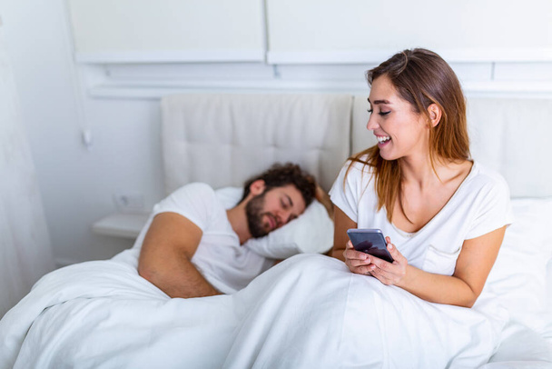 Cheating wife. Beautiful woman chatting privately on cellphone, hiding from her sleeping husband, empty space. Cheating Girlfriend Chatting On Phone While Boyfriend Sleeping In Bedroom At Night. - Photo, Image