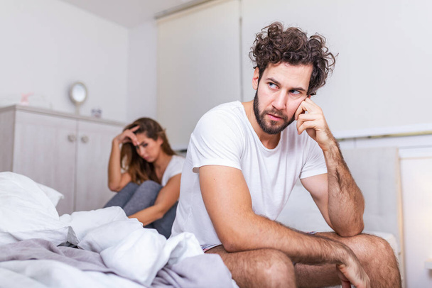 Young couple having a problem. Handsome man is leaning on his hand while sitting sadly on bed, woman is sitting in the background. Head full of thoughts when she is around - Photo, Image