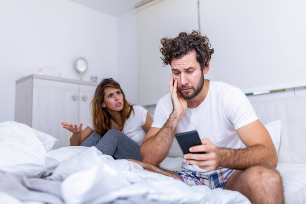 Το νεαρό ζευγάρι κάθεται στο κρεβάτι. Ο τύπος κοιτάει κάτι στο κινητό του. Το κορίτσι είναι προσβεβλημένο μαζί του. Τον κοιτάζει ενοχλημένη και απογοητευμένη από το αγόρι της στο τηλέφωνο. - Φωτογραφία, εικόνα