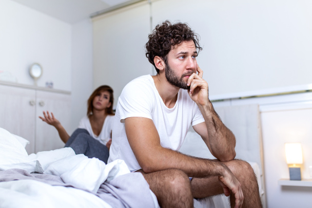 Beautiful girl and a frustrated man sitting in bed and not looking at each other. Upset couple ignoring each other. Worried man in tension at bed. Young couple angry with each other after a fight. - Photo, Image