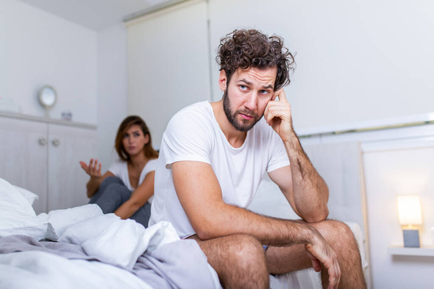 Young couple having a problem. Handsome man is leaning on his hand while sitting sadly on bed, woman is sitting in the background. Head full of thoughts when she is around - Foto, Imagem