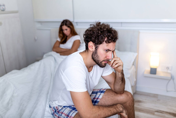 Beautiful girl and a frustrated man sitting in bed and not looking at each other. Upset couple ignoring each other. Worried man in tension at bed. Young couple angry with each other after a fight. - Φωτογραφία, εικόνα